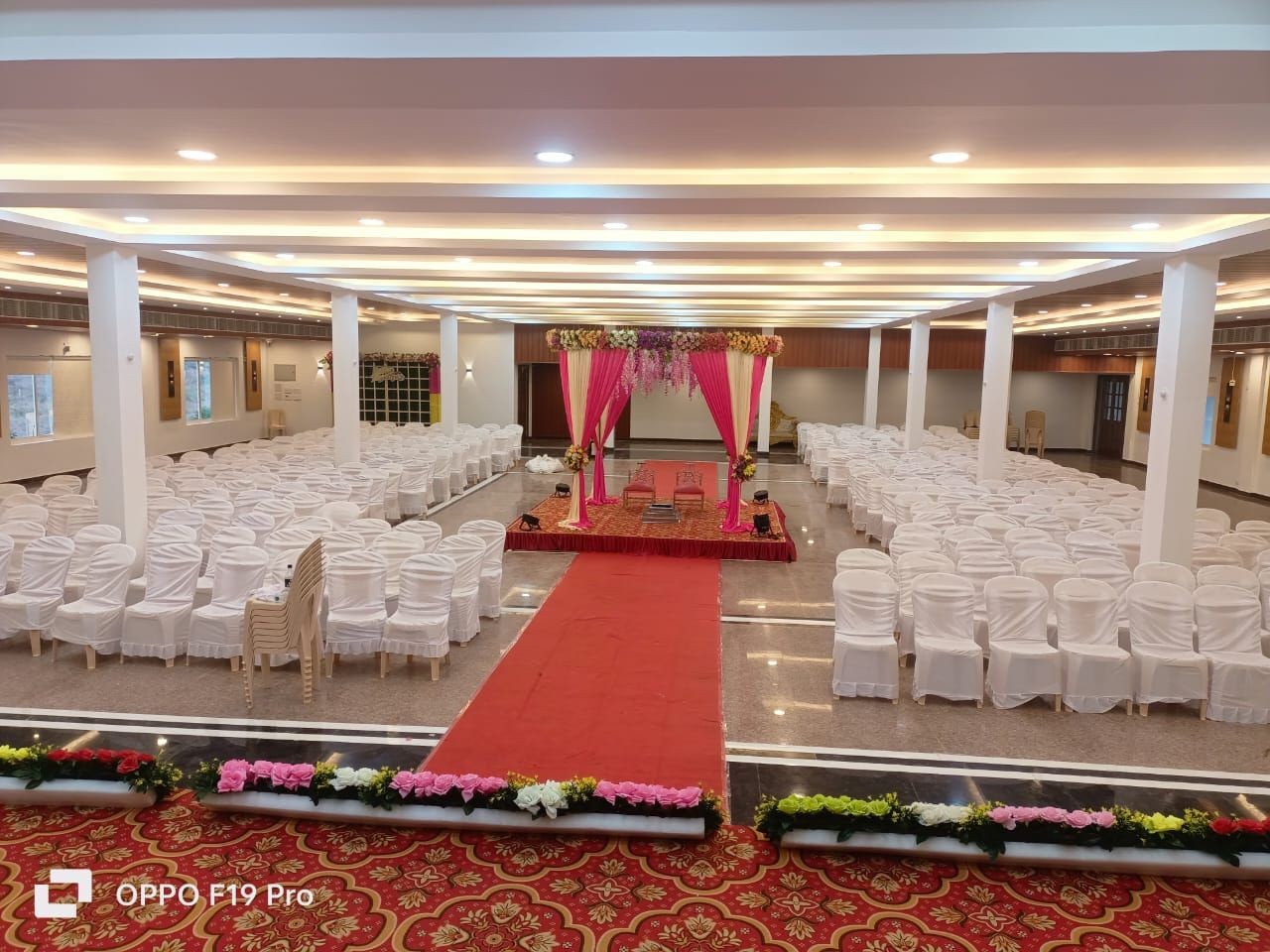 Empty indoor wedding hall with red carpet, decorated stage, and rows of white covered chairs.