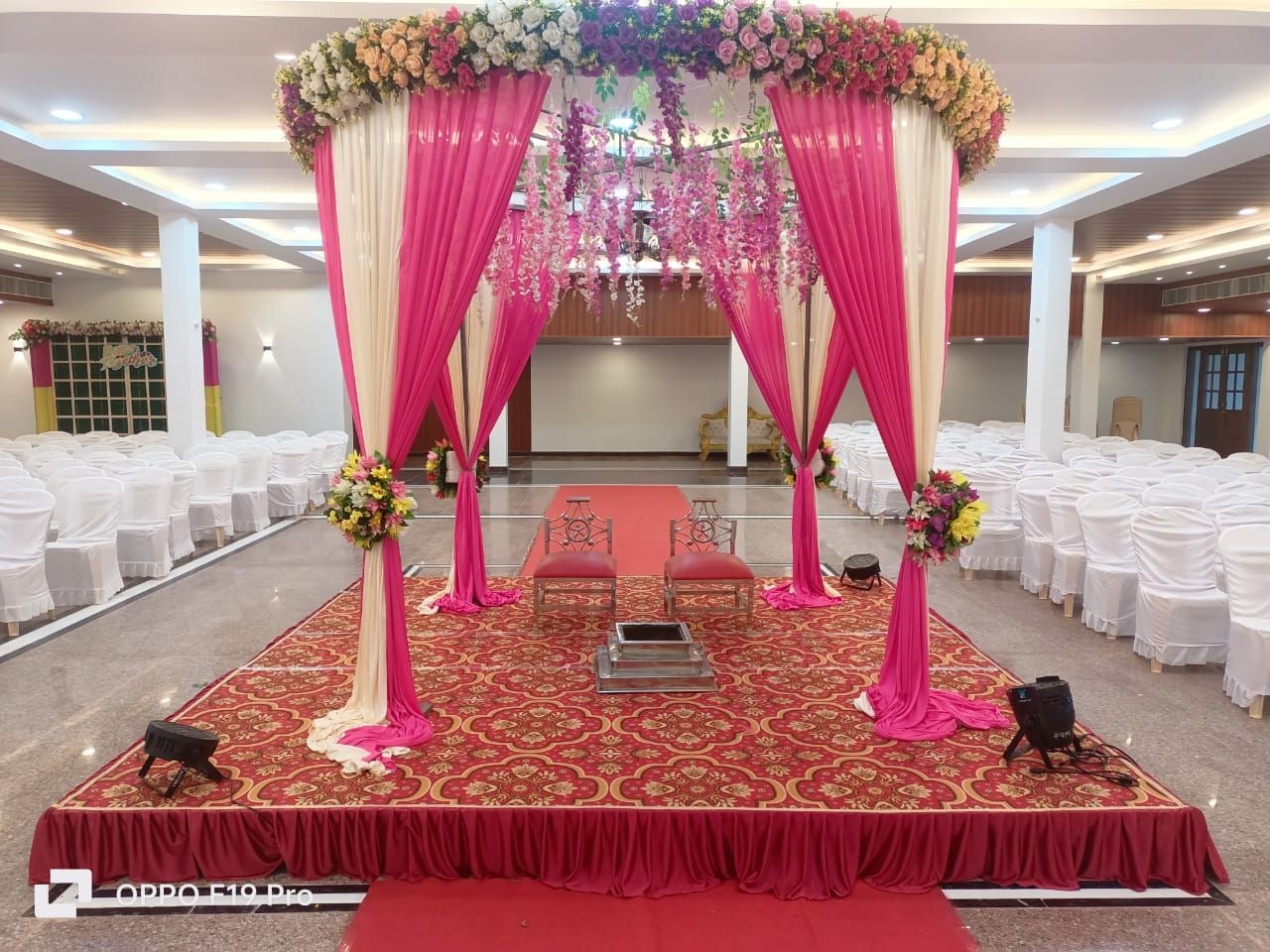 Indoor wedding venue setup with a decorated canopy, floral arrangements, and white chairs arranged in rows.